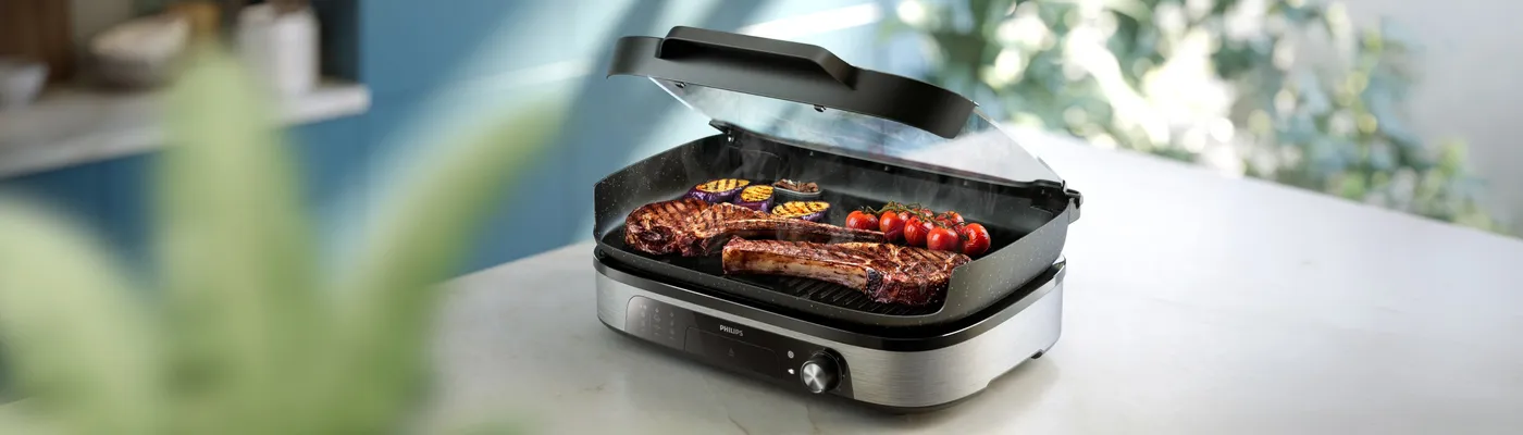 Philips electric grill with glass lid grilling steaks and vegetables on a kitchen countertop. High-temperature indoor grilling for juicy meat, grilled vegetables and everyday family meals. Philips electric grill with glass lid grilling steaks and vegetables on a kitchen countertop. High-temperature indoor grilling for juicy meat, grilled vegetables and everyday family meals.