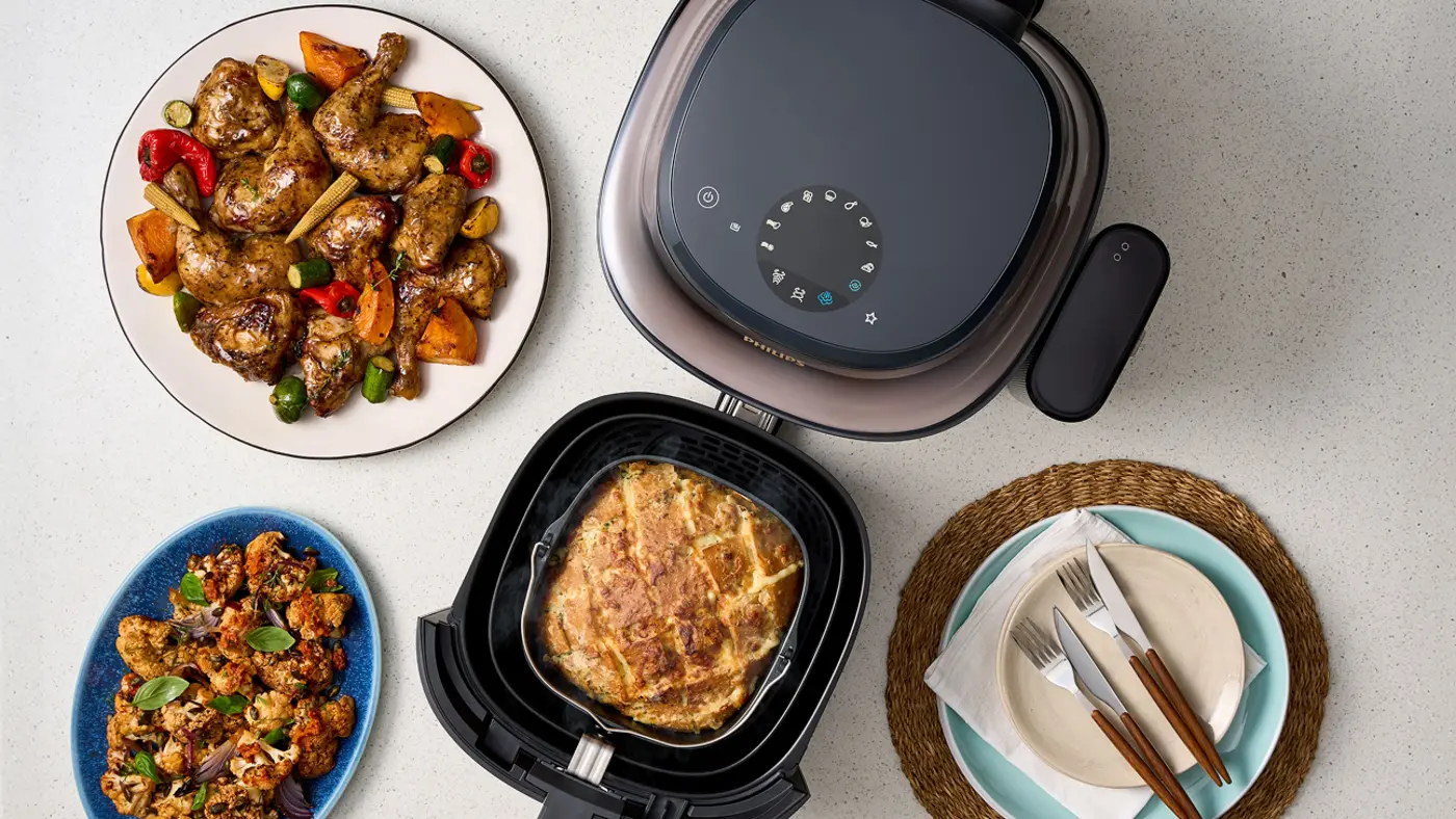 Philips Airfryer 5000 mit geöffnetem Korb, der einen gekochten Kuchen im Inneren zeigt. Um den Airfryer herum befinden sich Teller mit gekochten Hähnchenflügeln und geröstetem Gemüse, Besteck auf einer Serviette und eine moderne Küchenarbeitsplatte. Philips Airfryer 5000 mit geöffnetem Korb, der einen gekochten Kuchen im Inneren zeigt. Um den Airfryer herum befinden sich Teller mit gekochten Hähnchenflügeln und geröstetem Gemüse, Besteck auf einer Serviette und eine moderne Küchenarbeitsplatte.