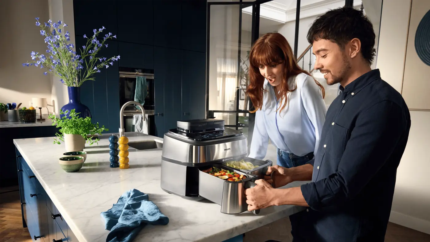 Paar genießt die Philips Kitchen Dual Basket Heißluftfritteuse, zeigt gefüllte Körbe mit Gemüse, Dampf steigt auf. Moderne Küche mit blauen Schränken und Marmorarbeitsplatte. Paar genießt die Philips Kitchen Dual Basket Heißluftfritteuse, zeigt gefüllte Körbe mit Gemüse, Dampf steigt auf. Moderne Küche mit blauen Schränken und Marmorarbeitsplatte.