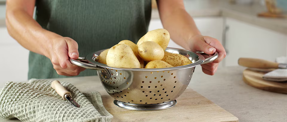 Henkilö pitelee metallisiivilää, joka on täynnä kuorimattomia perunoita. Siivilä on asetettu puisen leikkuulaudan päälle, ja sen vieressä on perunankuorija ja vihreä keittiöpyyhe. Henkilö pitelee metallisiivilää, joka on täynnä kuorimattomia perunoita. Siivilä on asetettu puisen leikkuulaudan päälle, ja sen vieressä on perunankuorija ja vihreä keittiöpyyhe.