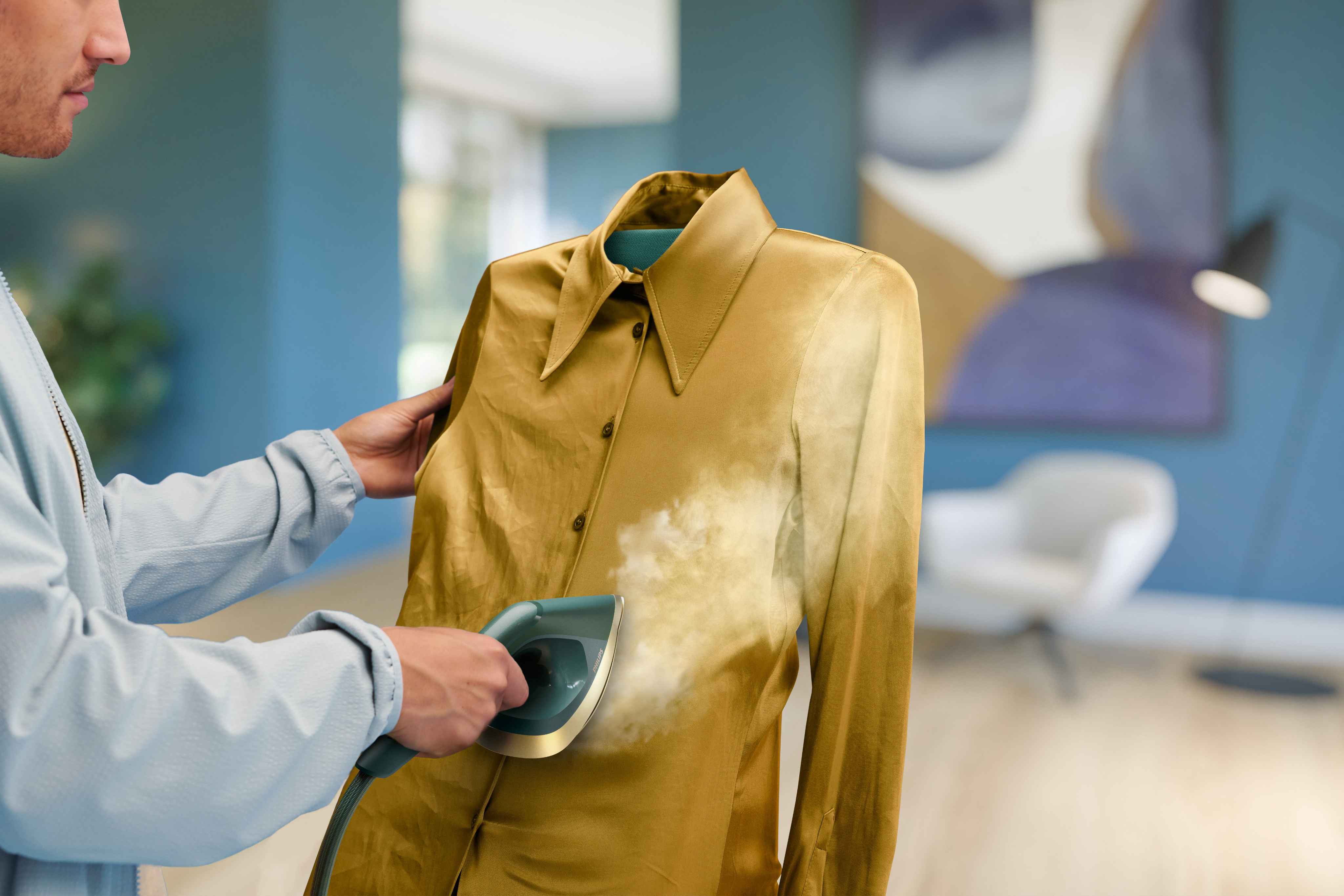 Soin du linge Philips All-in-One: personne utilisant le défroisseur sur un haut doré, montrant l'élimination immédiate des plis à la vapeur. Pièce moderne. Soin du linge Philips All-in-One: personne utilisant le défroisseur sur un haut doré, montrant l'élimination immédiate des plis à la vapeur. Pièce moderne.