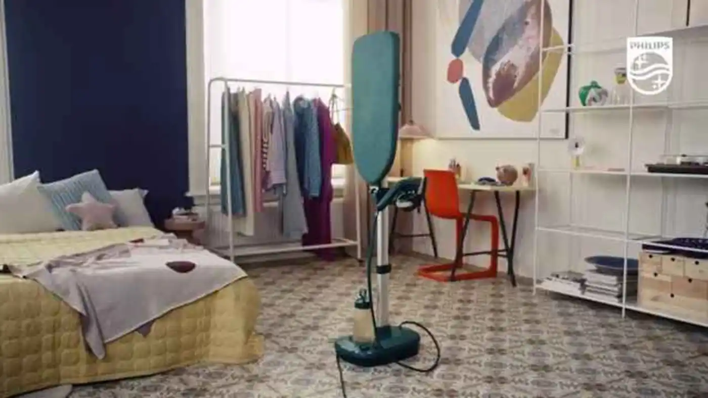 Philips All-in-One kledingverzorging: Een staande steamer met turquoise verticaal bord, zilveren accenten, in een stijlvolle kamer. Kledingrek en bed zichtbaar. Philips All-in-One kledingverzorging: Een staande steamer met turquoise verticaal bord, zilveren accenten, in een stijlvolle kamer. Kledingrek en bed zichtbaar.