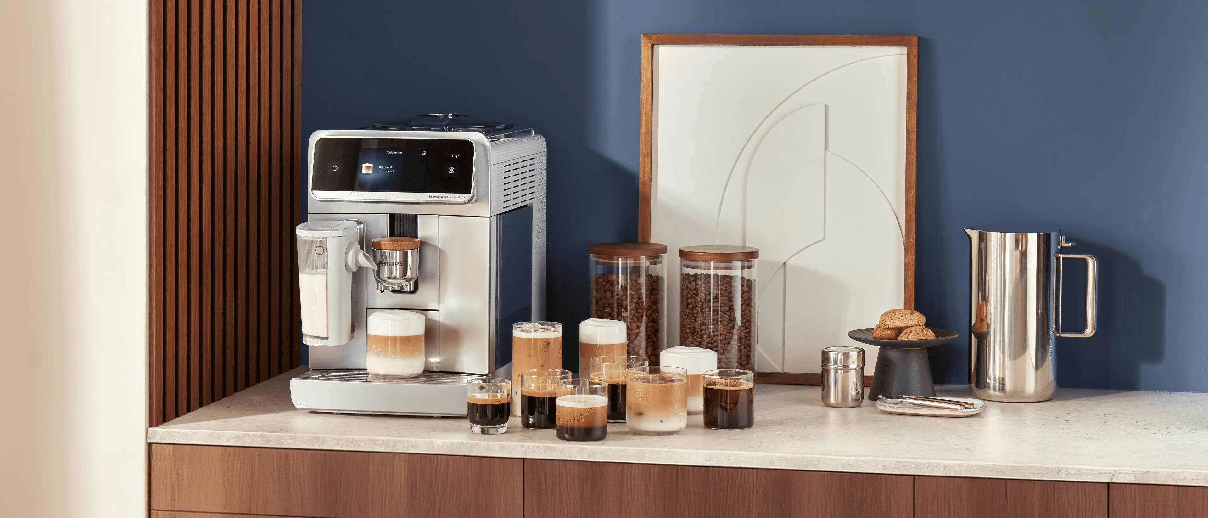 Automatyczny ekspres do kawy Philips 8000 LatteGo Pro Café Aromis na blacie kuchennym z kilkoma filiżankami napojów kawowych, słoikami ziaren i dzbankiem obok. Automatyczny ekspres do kawy Philips 8000 LatteGo Pro Café Aromis na blacie kuchennym z kilkoma filiżankami napojów kawowych, słoikami ziaren i dzbankiem obok.