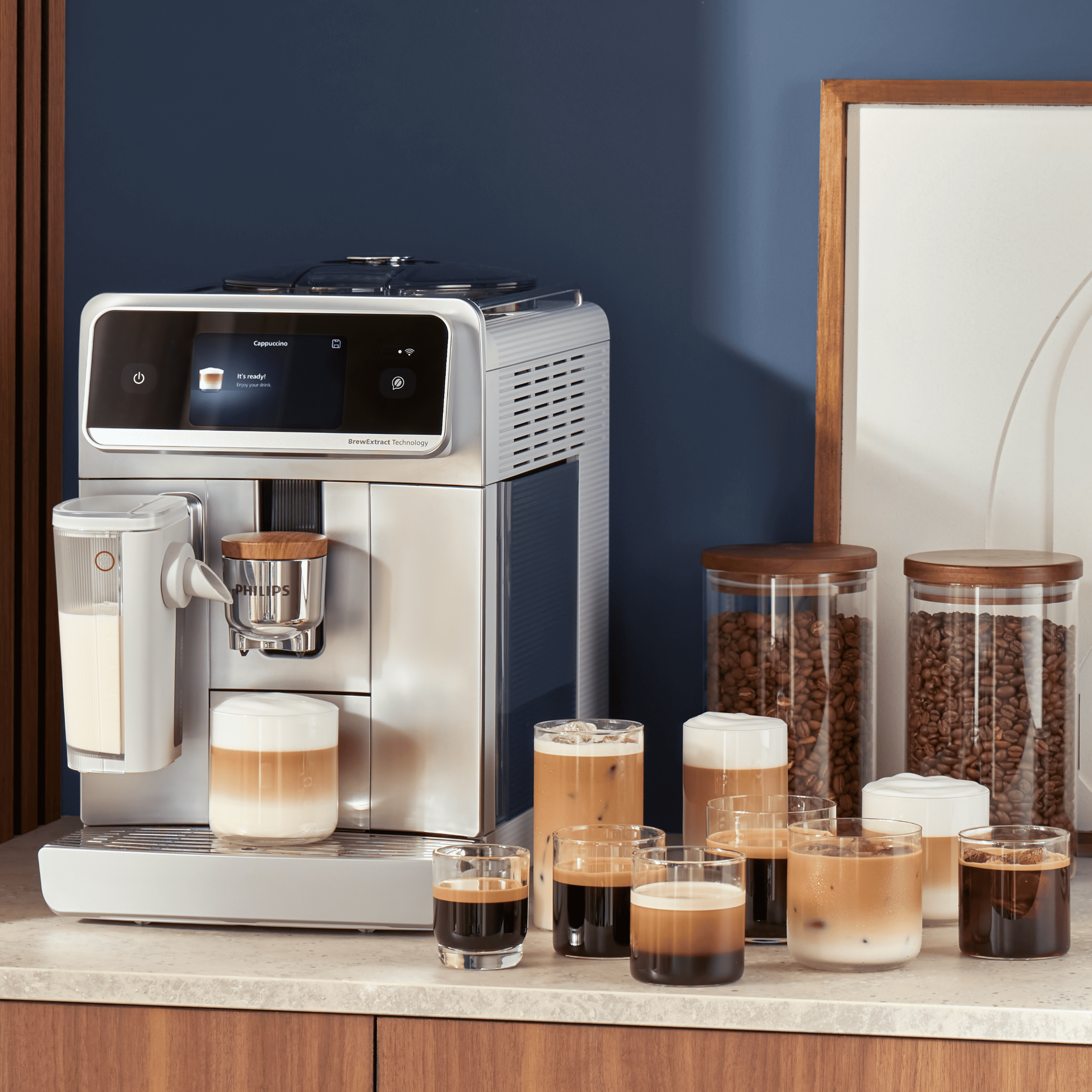 Une machine à expresso automatique Philips sur un comptoir de cuisine avec plusieurs tasses de boissons au café, des bocaux de grains et un pichet à côté. Une machine à expresso automatique Philips sur un comptoir de cuisine avec plusieurs tasses de boissons au café, des bocaux de grains et un pichet à côté.