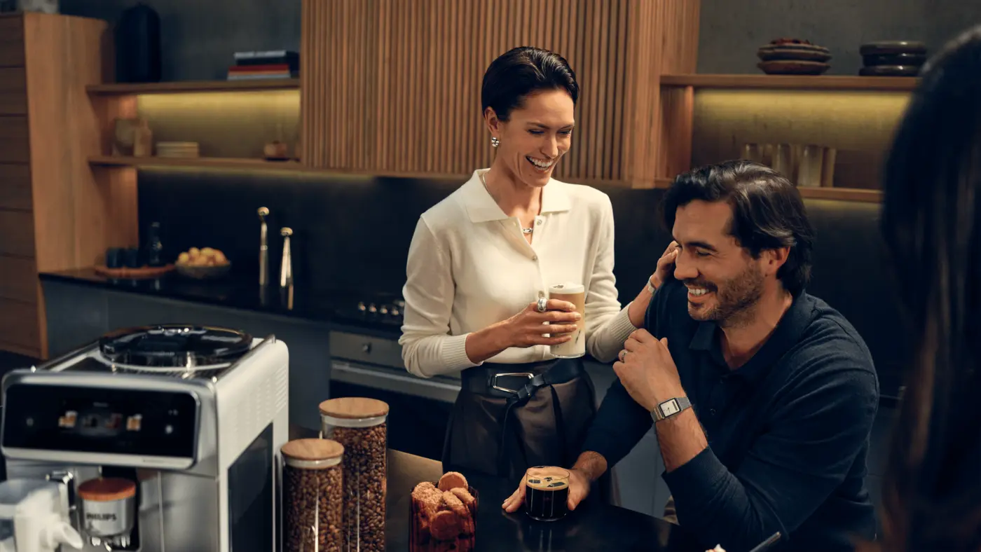 Philips Café Aromis 8000-seriens kaffemaskine på en moderne køkkenbordplade, mens et par nyder friskbrygget kaffe derhjemme Philips Café Aromis 8000-seriens kaffemaskine på en moderne køkkenbordplade, mens et par nyder friskbrygget kaffe derhjemme