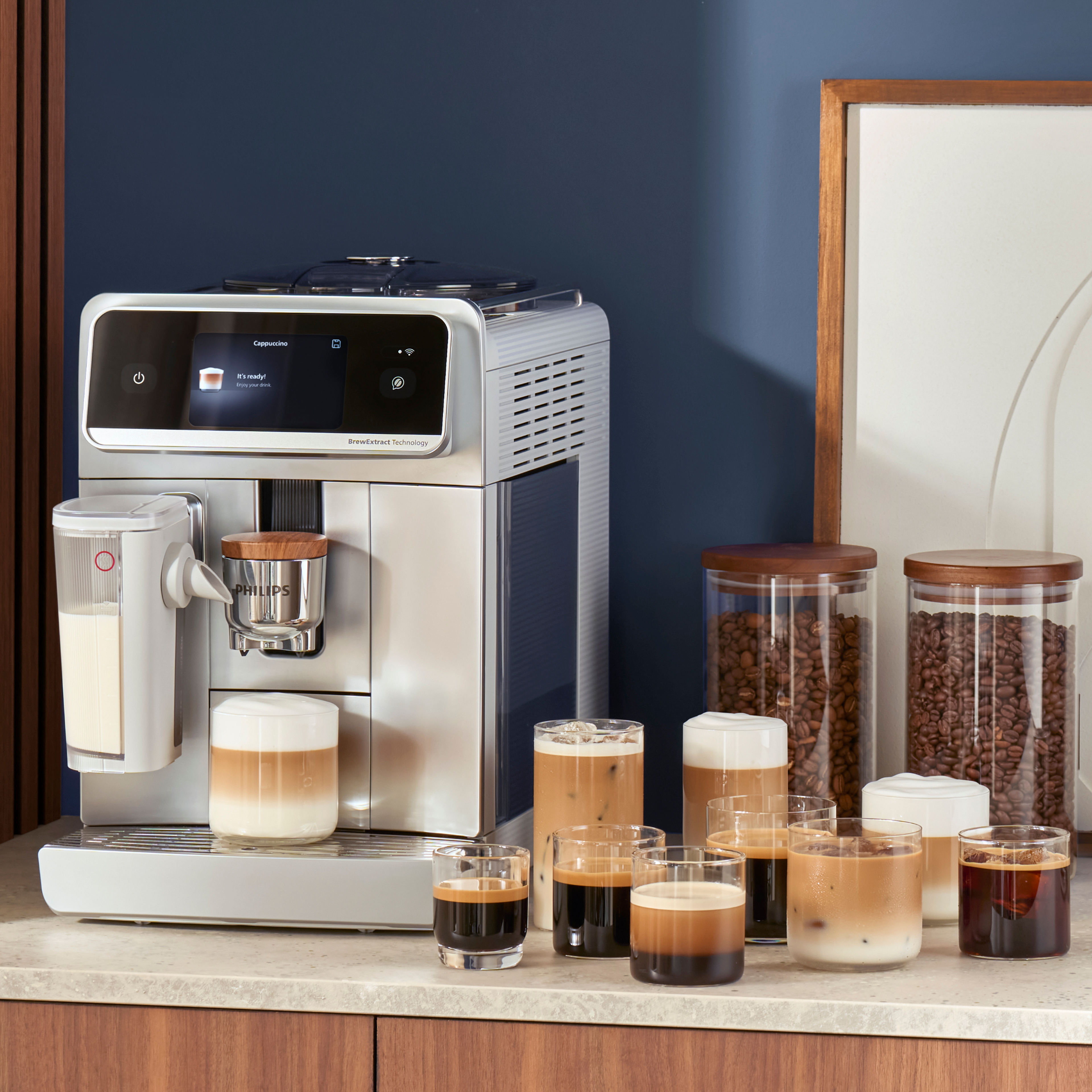 Ein Philips Kaffeevollautomat auf einer Küchenarbeitsplatte mit mehreren Tassen Kaffeegetränken, Gläsern mit Bohnen und einem Krug daneben. Ein Philips Kaffeevollautomat auf einer Küchenarbeitsplatte mit mehreren Tassen Kaffeegetränken, Gläsern mit Bohnen und einem Krug daneben.