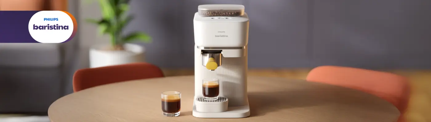 Philips Baristina coffee machine on a wooden table in a modern interior, dispensing espresso into a glass. A second espresso sits beside it, with a blurred plant and chairs in the background, creating a warm and stylish setting. Philips Baristina coffee machine on a wooden table in a modern interior, dispensing espresso into a glass. A second espresso sits beside it, with a blurred plant and chairs in the background, creating a warm and stylish setting.