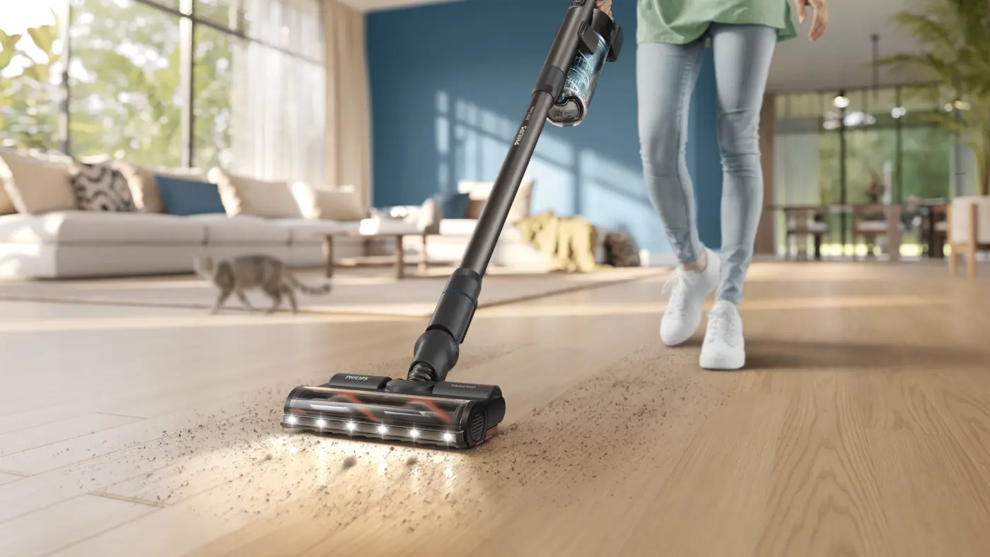Persona limpiando un suelo de madera con una aspiradora escoba Philips en un luminoso salón moderno con un gato en el fondo Persona limpiando un suelo de madera con una aspiradora escoba Philips en un luminoso salón moderno con un gato en el fondo