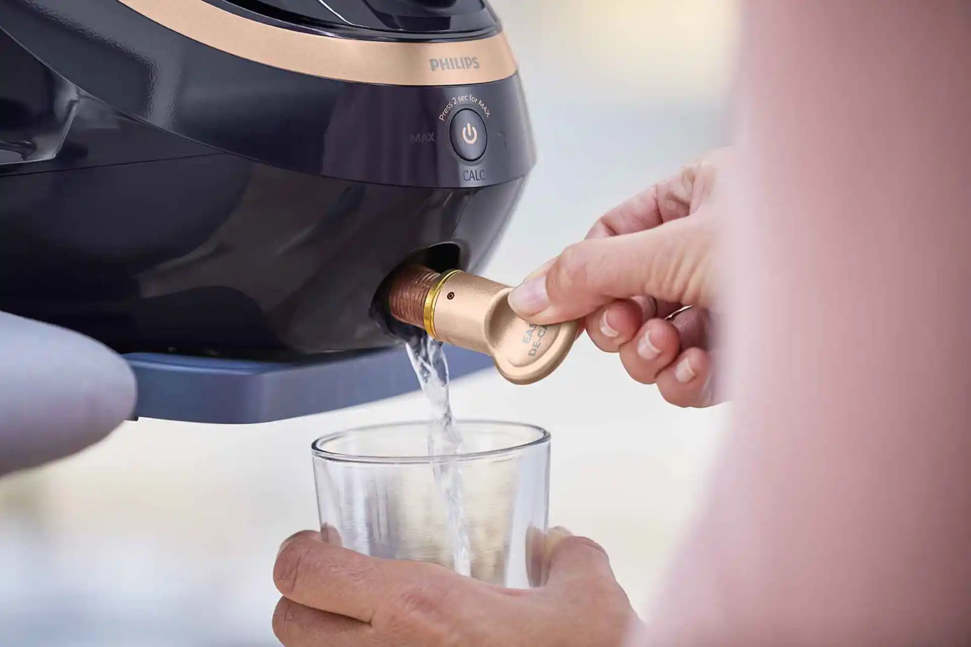 Nahaufnahme der Easy De-Calc Funktion eines Philips Garment Care Dampfglätters in Aktion. Wasser läuft in ein Glas und zeigt den einfachen Entkalkungsprozess, der die Lebensdauer des Dampfglätters verlängert. Nahaufnahme der Easy De-Calc Funktion eines Philips Garment Care Dampfglätters in Aktion. Wasser läuft in ein Glas und zeigt den einfachen Entkalkungsprozess, der die Lebensdauer des Dampfglätters verlängert.