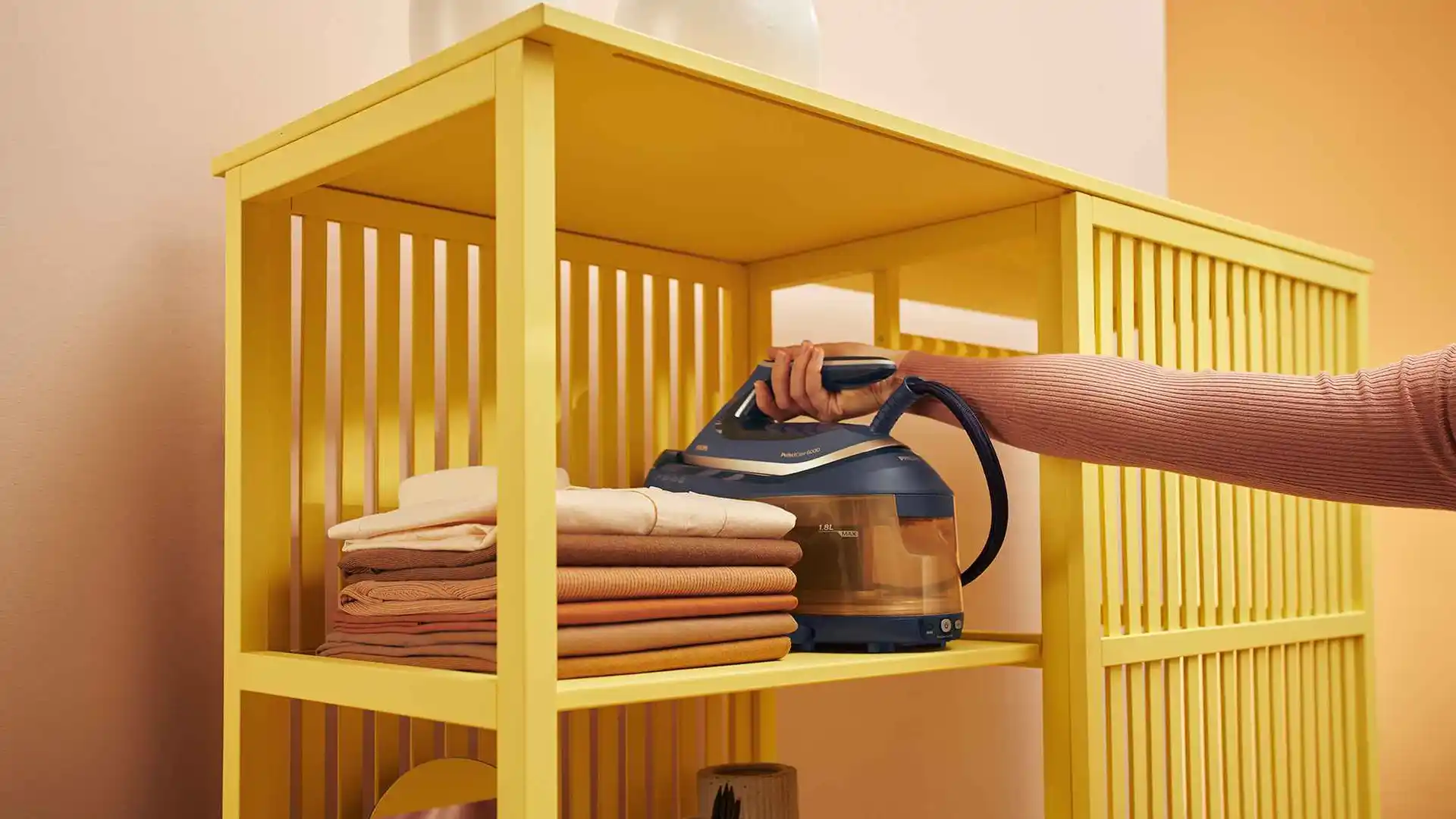 Der Philips Garment Care PerfectCare 6000 Serie Dampfgenerator steht auf einem Regal neben gefalteter Kleidung und präsentiert seinen 1,8-Liter-Wassertank und die einfache Bedienung. Der Philips Garment Care PerfectCare 6000 Serie Dampfgenerator steht auf einem Regal neben gefalteter Kleidung und präsentiert seinen 1,8-Liter-Wassertank und die einfache Bedienung.