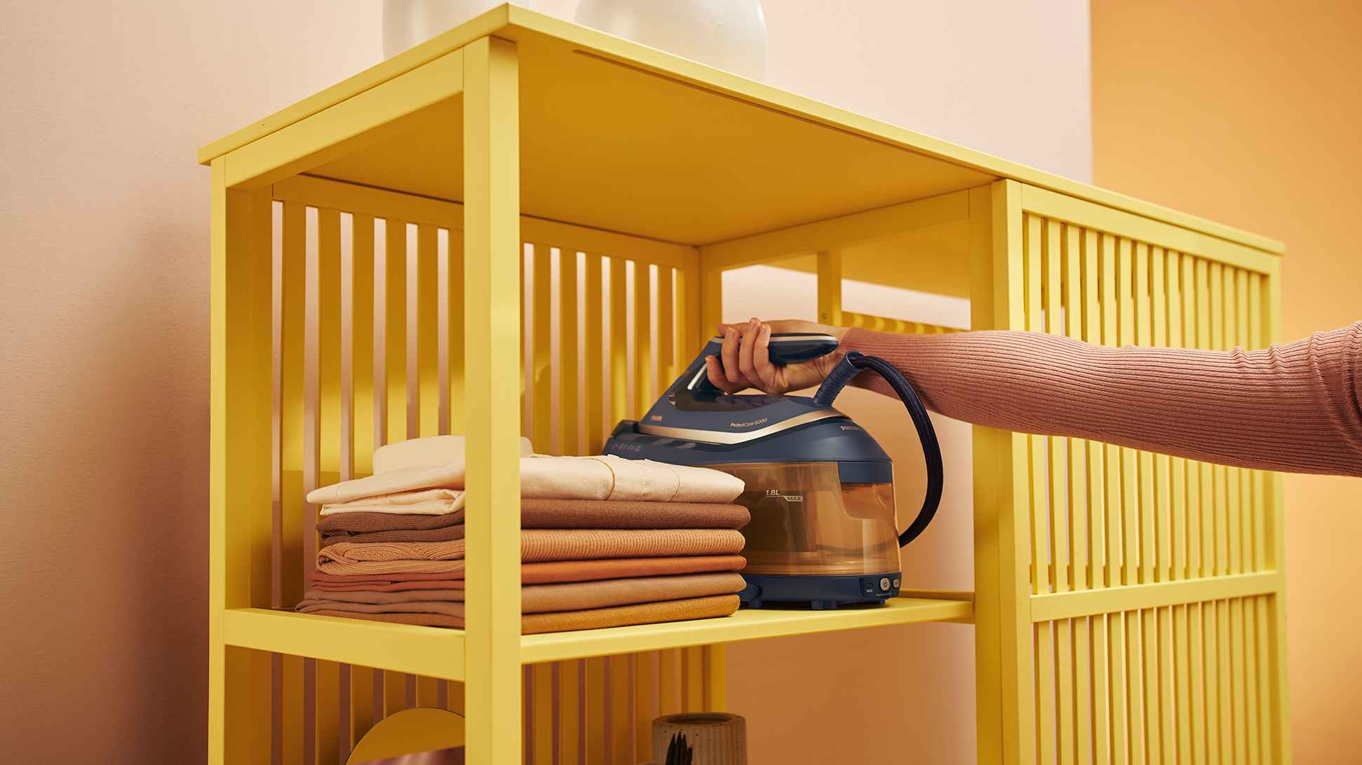 Philips Garment Care PerfectCare 6000 dampgenerator hviler på hylle ved siden av brettet tøy, og viser sin 1,8L vanntank og brukervennlighet. Philips Garment Care PerfectCare 6000 dampgenerator hviler på hylle ved siden av brettet tøy, og viser sin 1,8L vanntank og brukervennlighet.