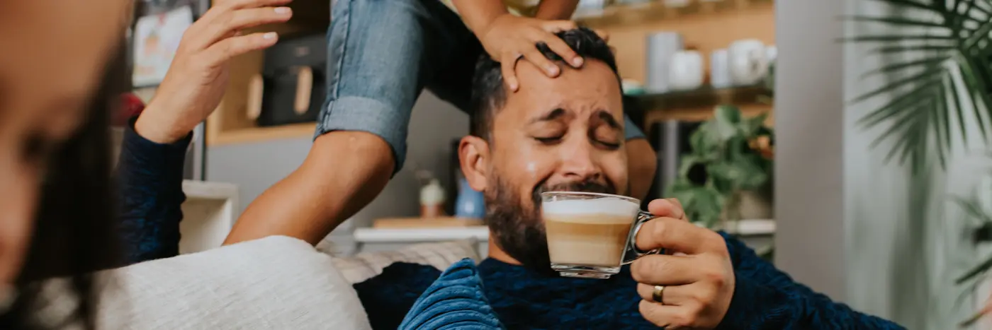 Homme souriant en buvant un café chez lui avec son enfant qui saute au-dessus de sa tête Homme souriant en buvant un café chez lui avec son enfant qui saute au-dessus de sa tête