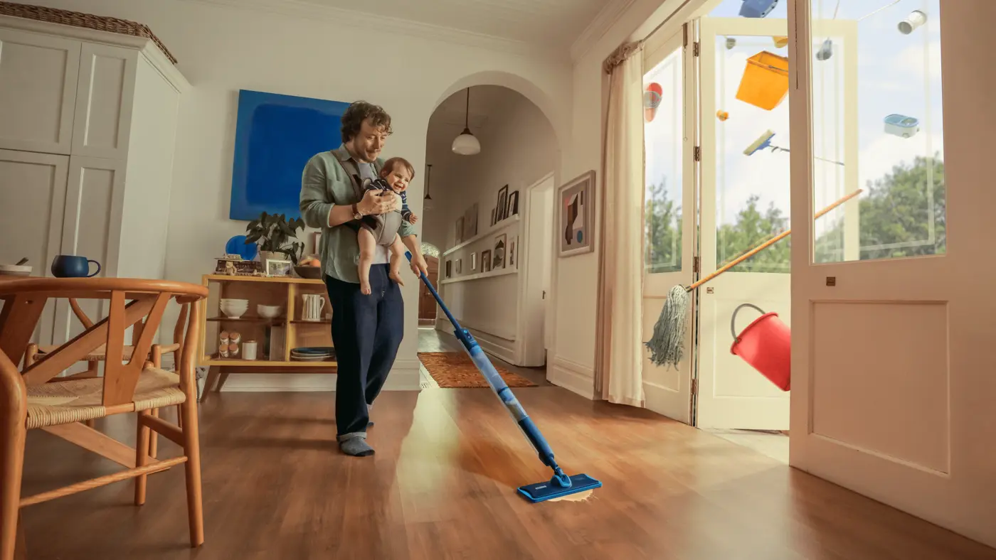 Eine Person wischt ein helles, modernes Wohnzimmer, während sie ein Baby im Arm hält; bunte Haushaltsgegenstände schweben durch die offenen Glastüren nach draußen. Eine Person wischt ein helles, modernes Wohnzimmer, während sie ein Baby im Arm hält; bunte Haushaltsgegenstände schweben durch die offenen Glastüren nach draußen.