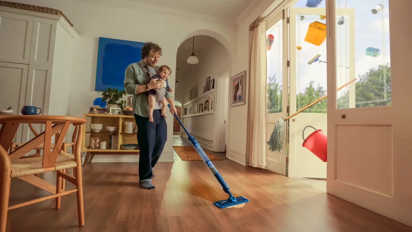 Člověk vytírá podlahu v jasném moderním obývacím pokoji, zatímco drží dítě v náručí; barevné předměty z domácnosti se vznáší ven skrz otevřené skleněné dveře. Člověk vytírá podlahu v jasném moderním obývacím pokoji, zatímco drží dítě v náručí; barevné předměty z domácnosti se vznáší ven skrz otevřené skleněné dveře.