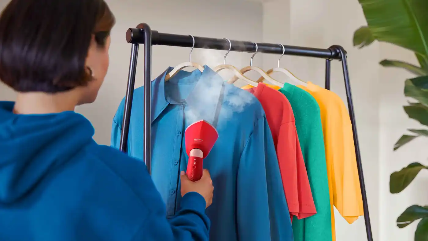 Persoon gebruikt een rode Philips Hand Steamer 5000 om snel kreukels uit een blauw overhemd op een rek te verwijderen. Ongeëvenaarde gebruikerservaring voor top kledingzorg. Persoon gebruikt een rode Philips Hand Steamer 5000 om snel kreukels uit een blauw overhemd op een rek te verwijderen. Ongeëvenaarde gebruikerservaring voor top kledingzorg.
