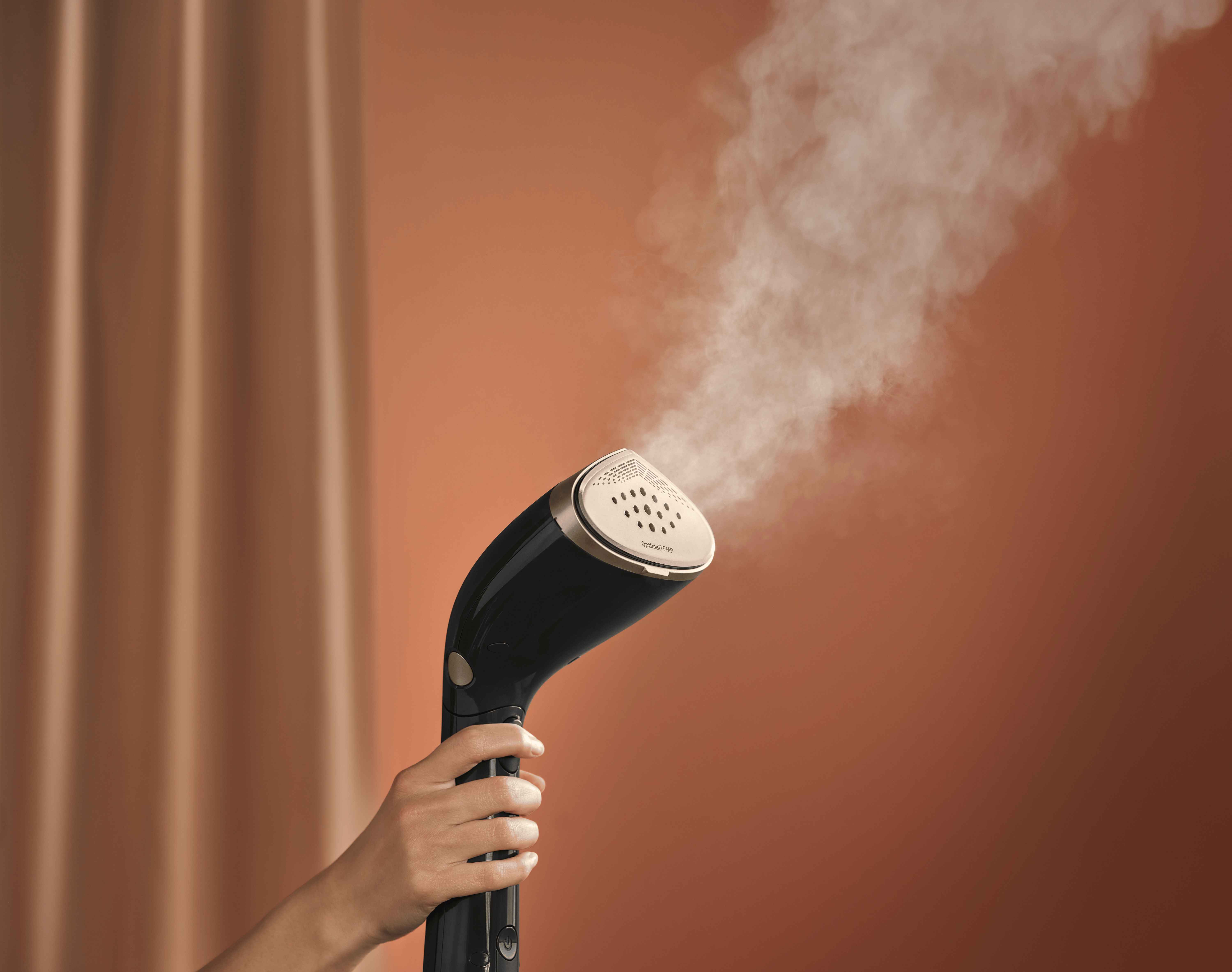 Eine Hand hält einen Philips Handheld Steamer, Teil der Philips Garment Care Linie, der mühelos Dampf abgibt. Hebt das schwarz-goldene Design hervor und betont die innovative Textilpflege. Eine Hand hält einen Philips Handheld Steamer, Teil der Philips Garment Care Linie, der mühelos Dampf abgibt. Hebt das schwarz-goldene Design hervor und betont die innovative Textilpflege.