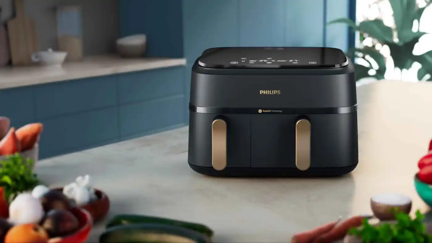 De Philips Dual Basket airfryer in donkere uitvoering met gouden handgrepen op aanrecht, toont capaciteit & technologie voor olievrije resultaten. De Philips Dual Basket airfryer in donkere uitvoering met gouden handgrepen op aanrecht, toont capaciteit & technologie voor olievrije resultaten.