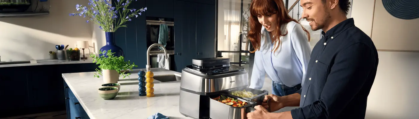Pár élvezi a Philips dupla kosaras airfryer használatát, zöldségekkel teli dupla kosarak, gőz száll fel. Modern konyha kék szekrényekkel és márványlappal. Pár élvezi a Philips dupla kosaras airfryer használatát, zöldségekkel teli dupla kosarak, gőz száll fel. Modern konyha kék szekrényekkel és márványlappal.