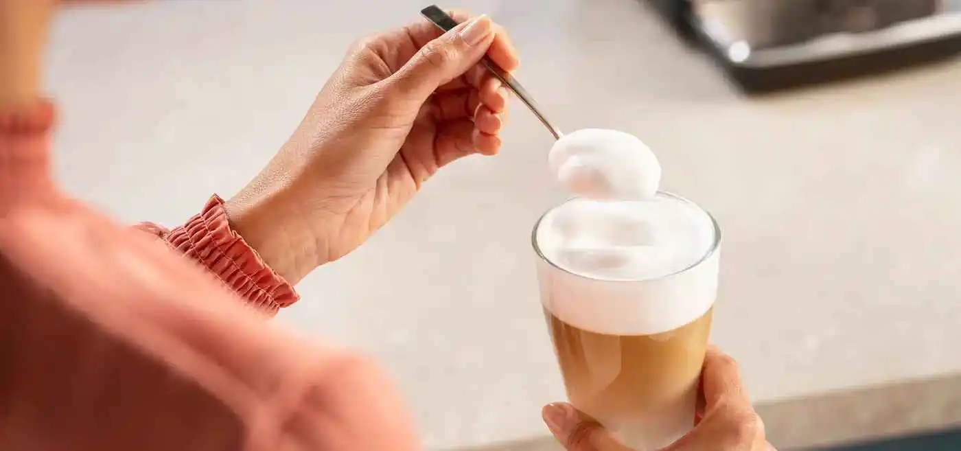 Una persona añade espuma de leche densa a un café con leche Philips LatteGo en capas con cuchara, resaltando la espuma perfecta para bebidas con calidad de cafetería en casa. Una persona añade espuma de leche densa a un café con leche Philips LatteGo en capas con cuchara, resaltando la espuma perfecta para bebidas con calidad de cafetería en casa.