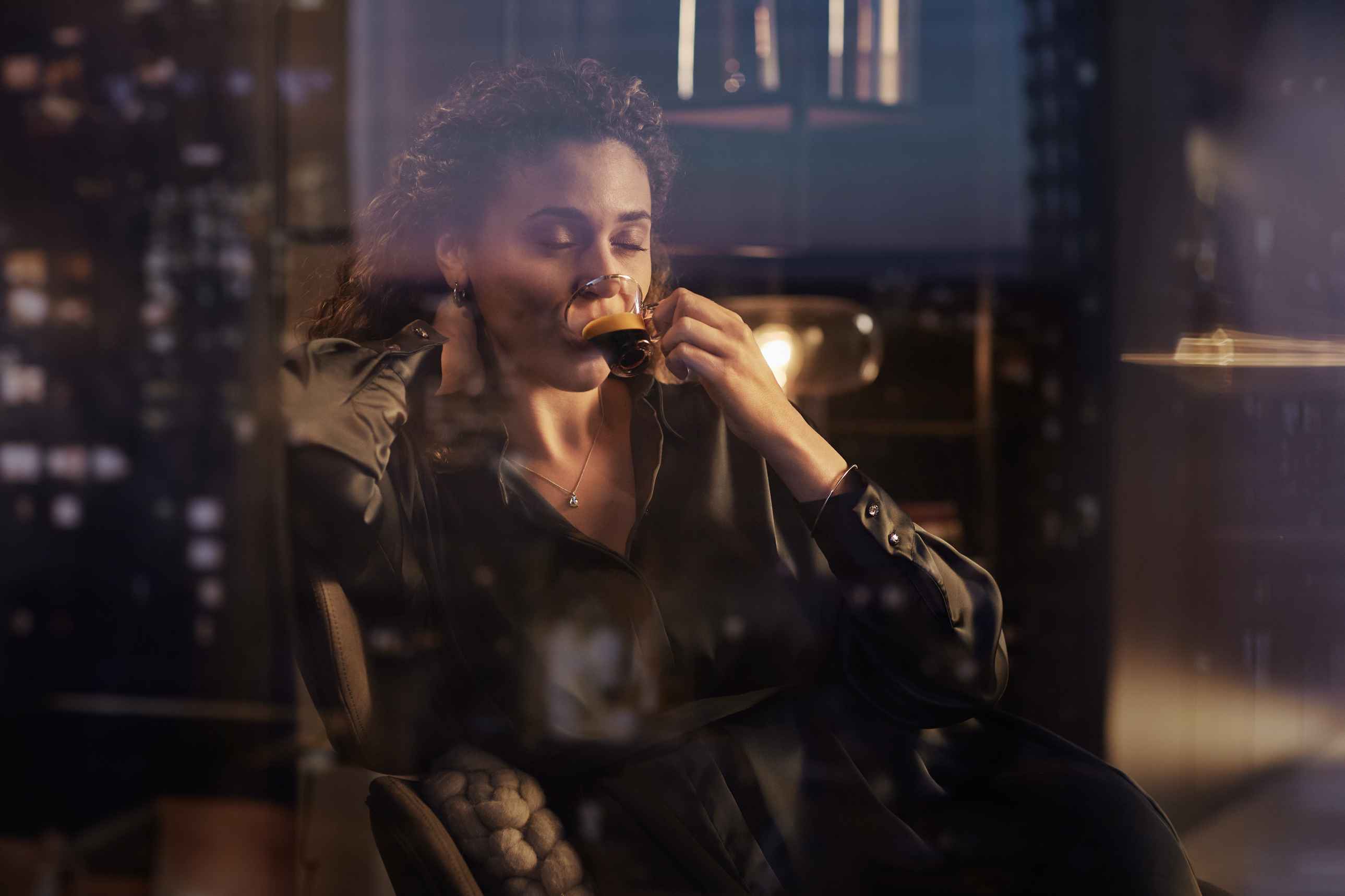 Une femme ferme les yeux en savourant une tasse de café noir et riche dans une tasse en verre transparente, sur fond de ville flou. Assise dans un fauteuil marron, elle porte une chemise verte soyeuse de la ligne Philips Lor. Une femme ferme les yeux en savourant une tasse de café noir et riche dans une tasse en verre transparente, sur fond de ville flou. Assise dans un fauteuil marron, elle porte une chemise verte soyeuse de la ligne Philips Lor.