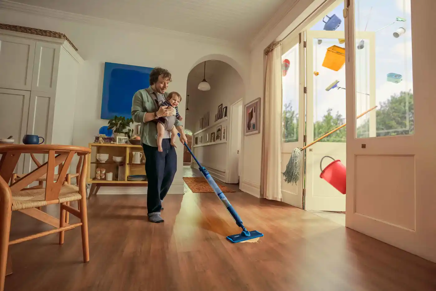Publicité Philips OneUp Entretien des sols : Père tenant un bébé tout en passant la serpillière, avec des produits de nettoyage sortant des portes ouvertes. Publicité Philips OneUp Entretien des sols : Père tenant un bébé tout en passant la serpillière, avec des produits de nettoyage sortant des portes ouvertes.