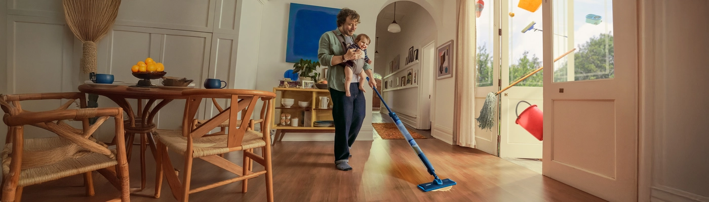 Egy személy egy csecsemőt tartva felmos egy világos, modern nappalit; színes háztartási cikkek lebegnek kint a nyitott üvegajtókon keresztül. Egy személy egy csecsemőt tartva felmos egy világos, modern nappalit; színes háztartási cikkek lebegnek kint a nyitott üvegajtókon keresztül.