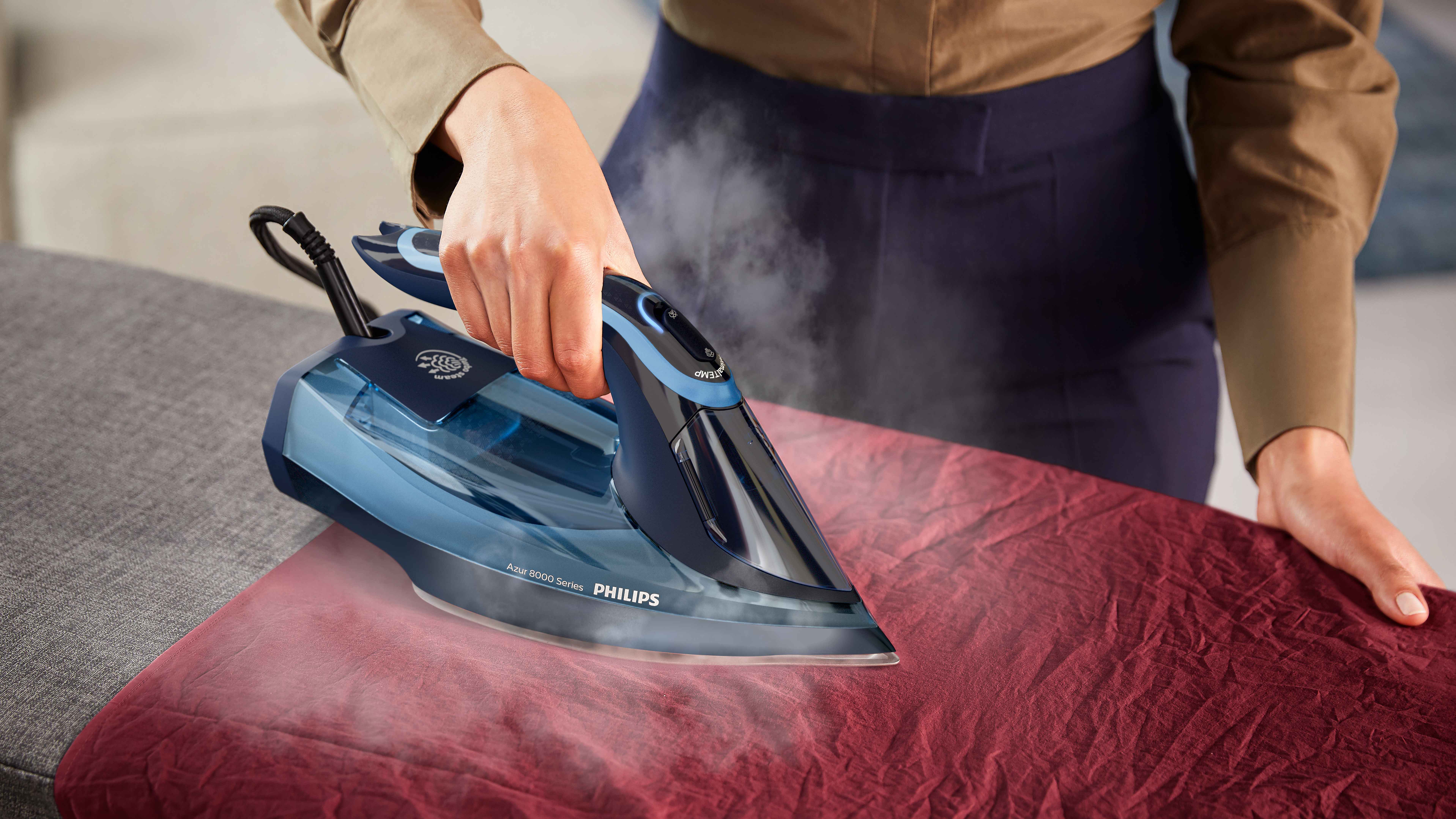 Philips Dampfbügeleisen: Person bügelt roten Stoff mit dem blau-schwarzen Azur 8000, Dampf tritt aus, demonstriert Präzision. Philips Dampfbügeleisen: Person bügelt roten Stoff mit dem blau-schwarzen Azur 8000, Dampf tritt aus, demonstriert Präzision.