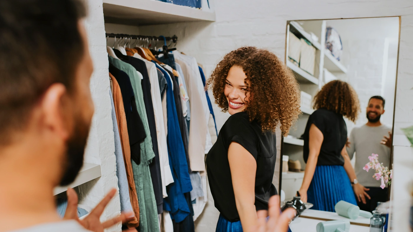A woman stands in front of a mirror in a dressing room with clothes on hangers; another person, seen from behind, gestures with her hands. A woman stands in front of a mirror in a dressing room with clothes on hangers; another person, seen from behind, gestures with her hands.