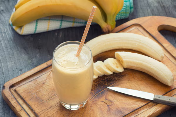 Un smoothie răcoritor de banane într-un pahar, așezat pe o tăietură cu un cuțit și banane coapte lângă el. Un smoothie răcoritor de banane într-un pahar, așezat pe o tăietură cu un cuțit și banane coapte lângă el.