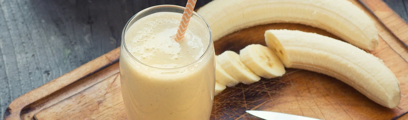 Un pahar cu smoothie de banane pe o tăietură cu un cuțit, înconjurat de banane proaspete Un pahar cu smoothie de banane pe o tăietură cu un cuțit, înconjurat de banane proaspete