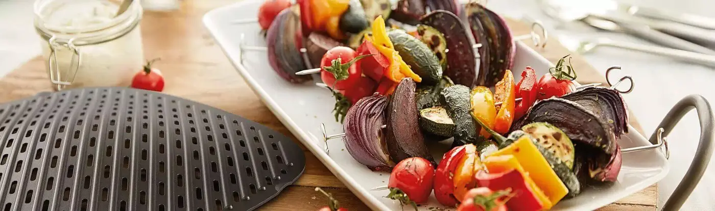 Ein Teller mit buntem Grillgemüse steht neben einem Grill Ein Teller mit buntem Grillgemüse steht neben einem Grill