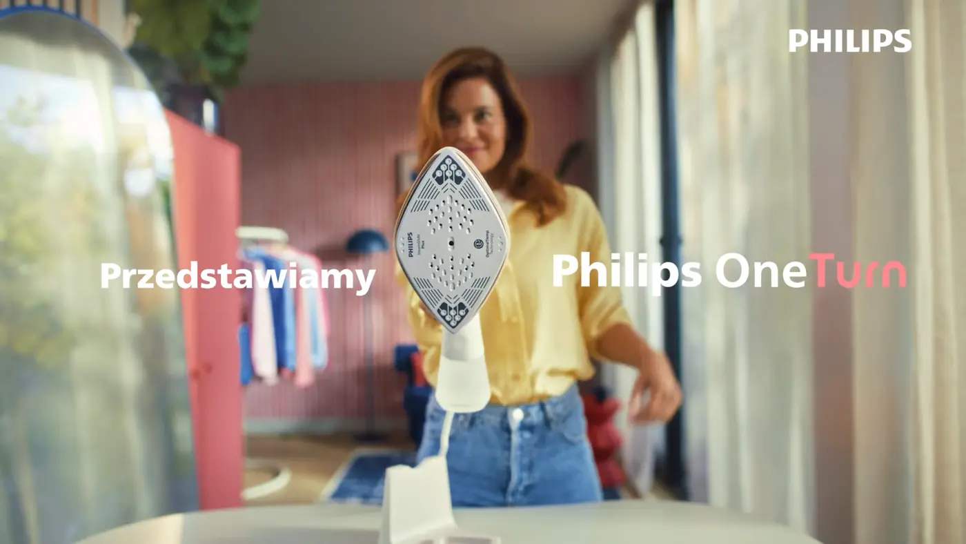 Żelazko i parownica do ubrań Philips OneTurn trzymane w pozycji pionowej na desce do prasowania przez kobietę w jasnym wnętrzu domu, z napisem „Przedstawiamy Philips OneTurn” i logo Philips na ekranie. Żelazko i parownica do ubrań Philips OneTurn trzymane w pozycji pionowej na desce do prasowania przez kobietę w jasnym wnętrzu domu, z napisem „Przedstawiamy Philips OneTurn” i logo Philips na ekranie.