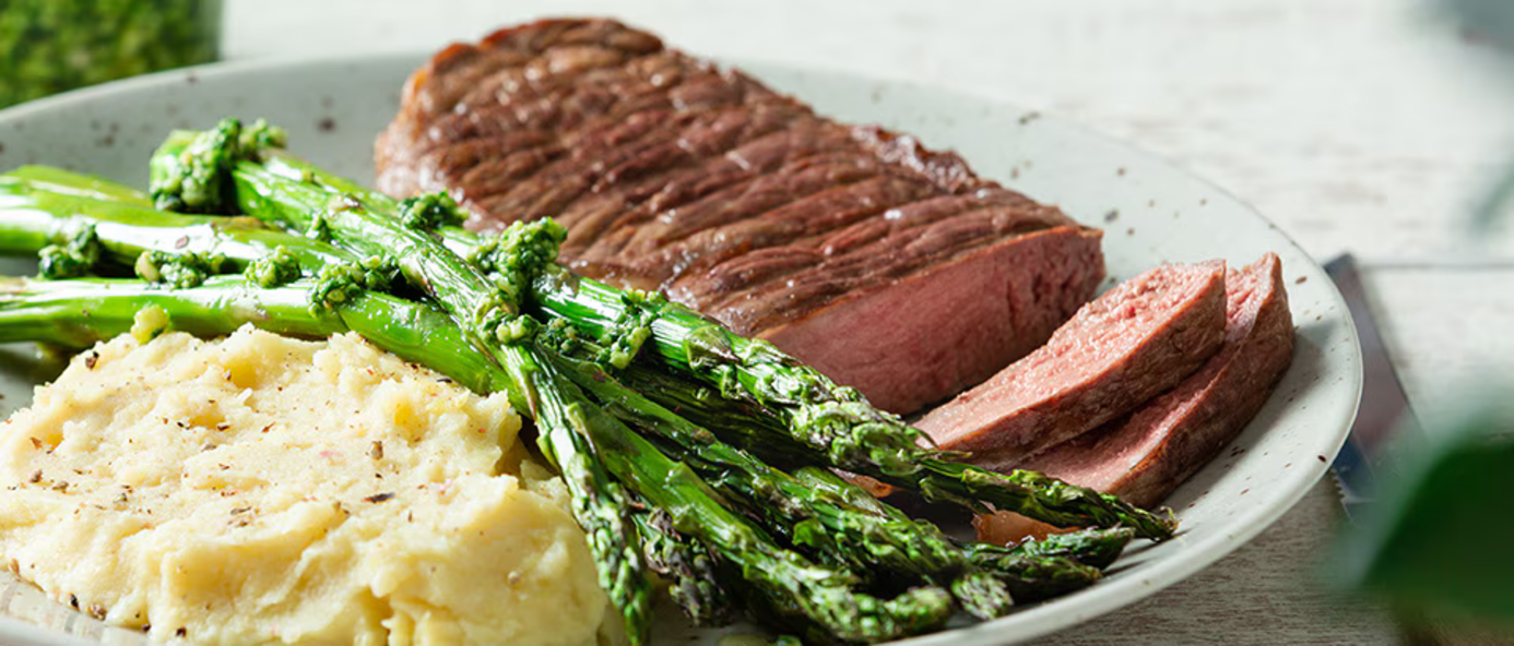 A plate of sliced, perfectly cooked steak served with roasted asparagus topped with herb pesto and creamy mashed potatoes. The dish is arranged on a white plate with a rustic, fresh presentation. A plate of sliced, perfectly cooked steak served with roasted asparagus topped with herb pesto and creamy mashed potatoes. The dish is arranged on a white plate with a rustic, fresh presentation.